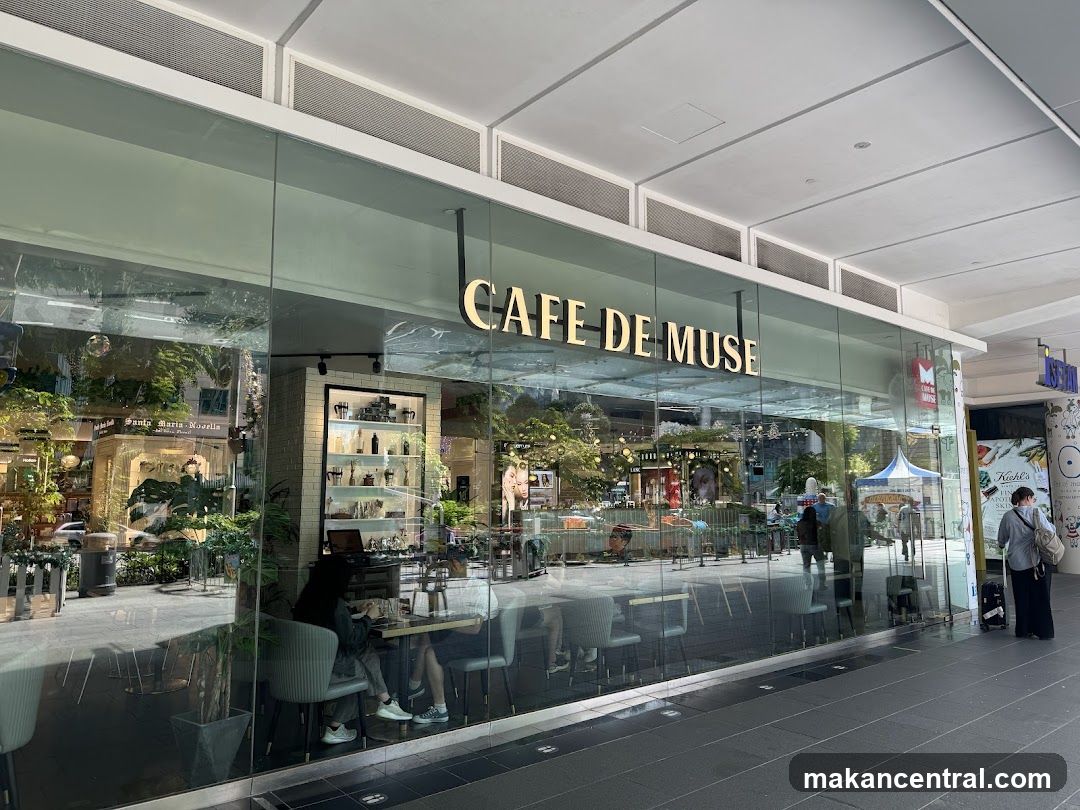 Modern glass facade with gold lettering 'CAFE DE MUSE' above entrance. Interior shows seated patrons, greenery, shelves with products, and a bright, airy ambiance. Reflections of the surrounding mall are visible on glass. A person stands outside near the right edge.