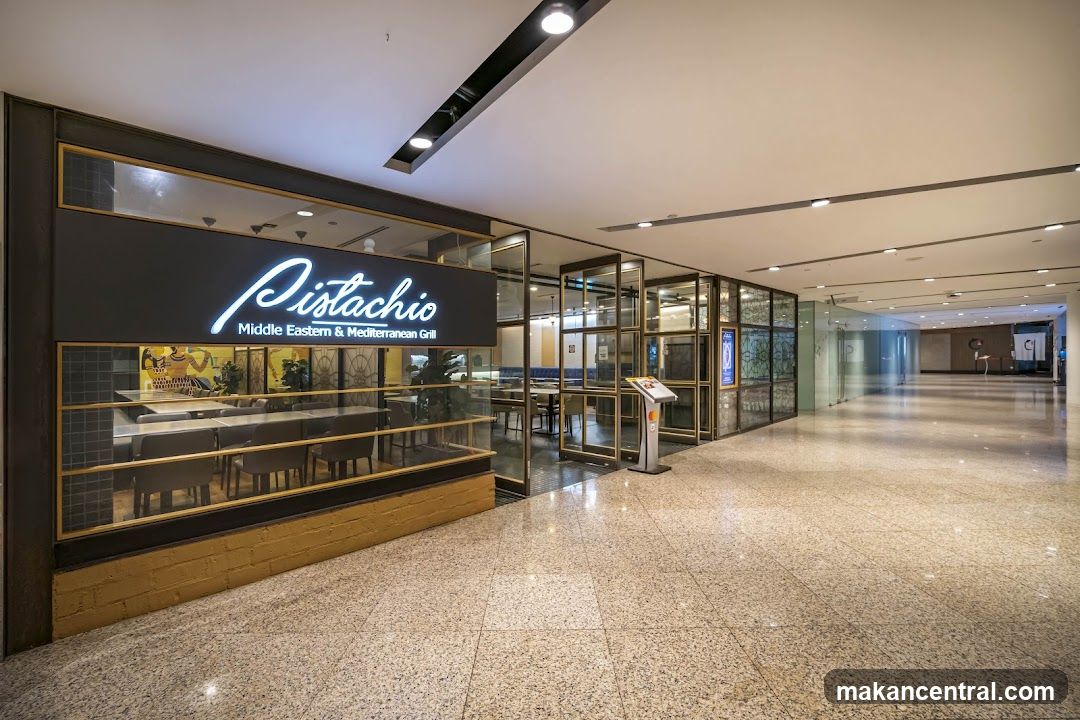 Modern storefront with large glass windows framed in gold trim, featuring a prominent neon sign reading 'Pistachio Middle Eastern & Mediterranean Grill'. The entrance has glass doors and a visible interior dining area with tables and chairs. A small kiosk or information stand is positioned near the doorway. The floor is polished stone, and ceiling lights illuminate the space.