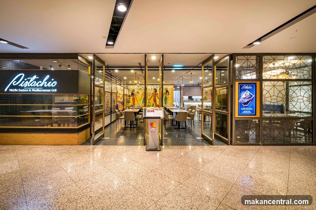 Storefront with illuminated sign on dark panel above glass entrance; gold-trimmed doors open to dining area with tables, chairs, and decorative murals; right side features ornate patterned glass panels and a digital display screen showing promotional content.