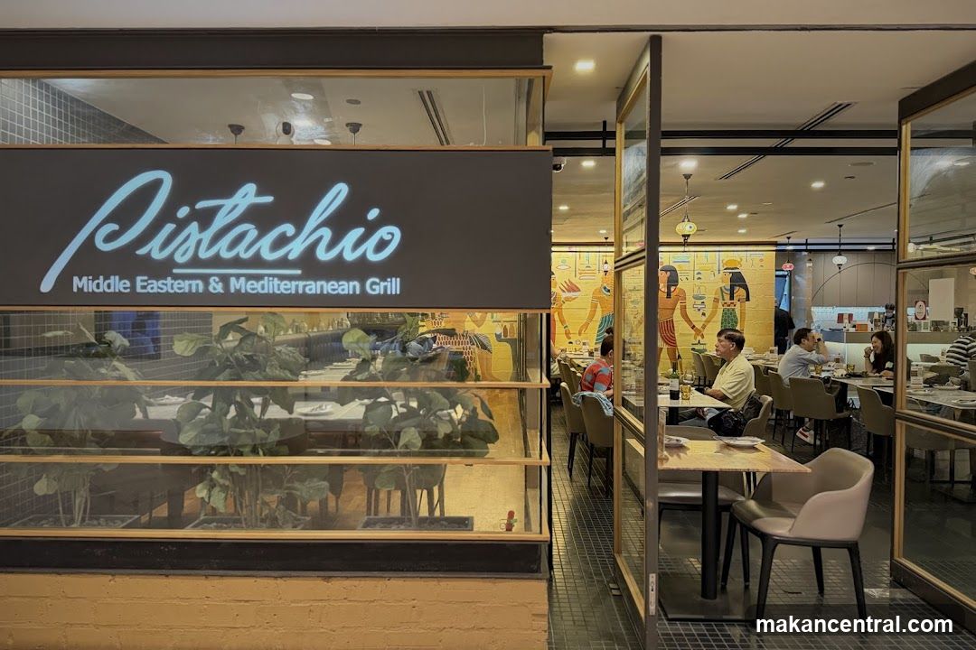 Exterior storefront with illuminated sign reading 'Pistachio Middle Eastern & Mediterranean Grill'. Glass windows display indoor dining area with plants in foreground. Visible interior includes patrons seated at tables, Egyptian-themed wall art, and modern lighting.
