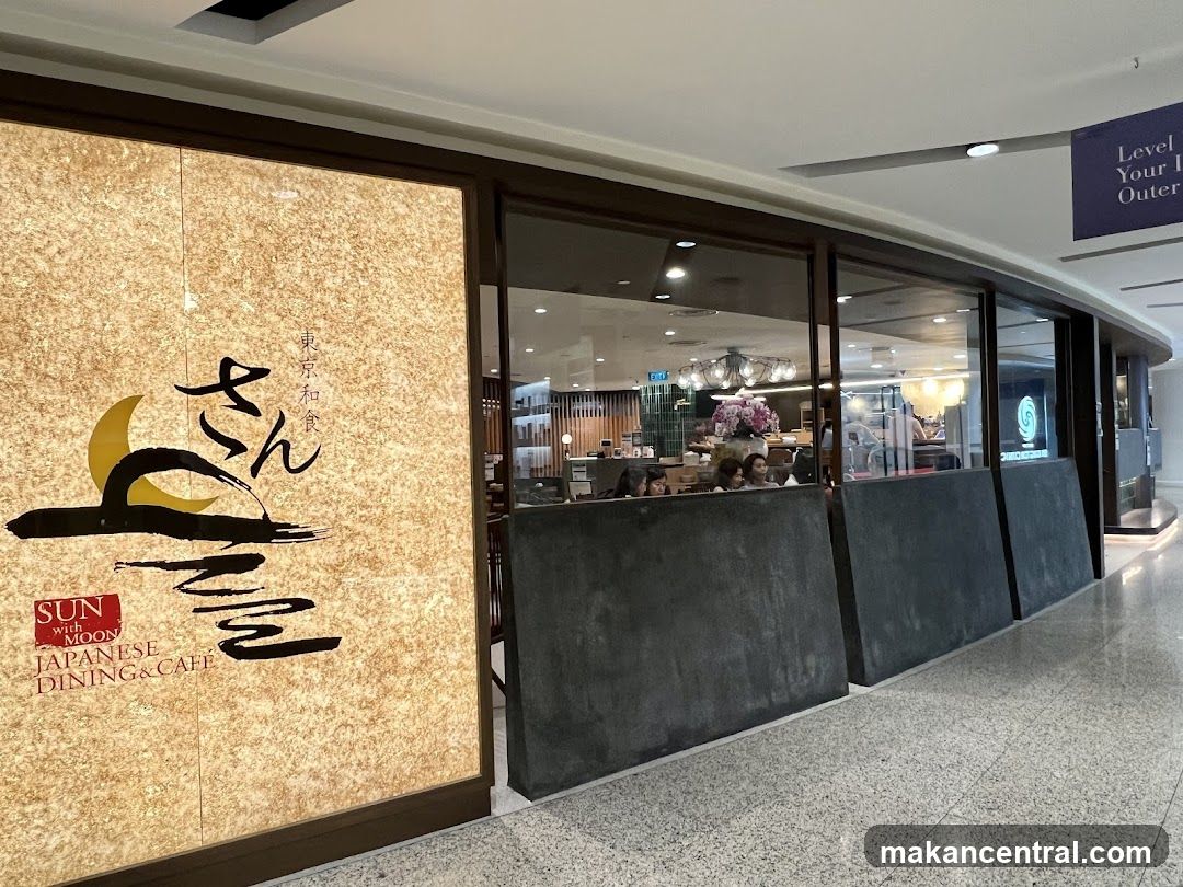 Exterior storefront with large textured wall featuring logo and name in stylized calligraphy alongside English text 'SUN with MOON JAPANESE DINING&CAFE'. Glass windows reveal interior seating area with patrons, chandeliers, and floral arrangements. Dark gray counter fronts the entrance. Above right, a blue sign reads 'Level 1 Your Outer' partially visible.