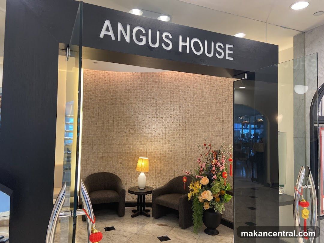 Modern glass storefront with black frame and white "ANGUS HOUSE" signage above entrance. Interior features beige tiled wall, two dark armchairs, a small table with a lit lamp, and a large floral arrangement in a black pot. Red tassels hang from door handles. Reflections show interior dining area.
