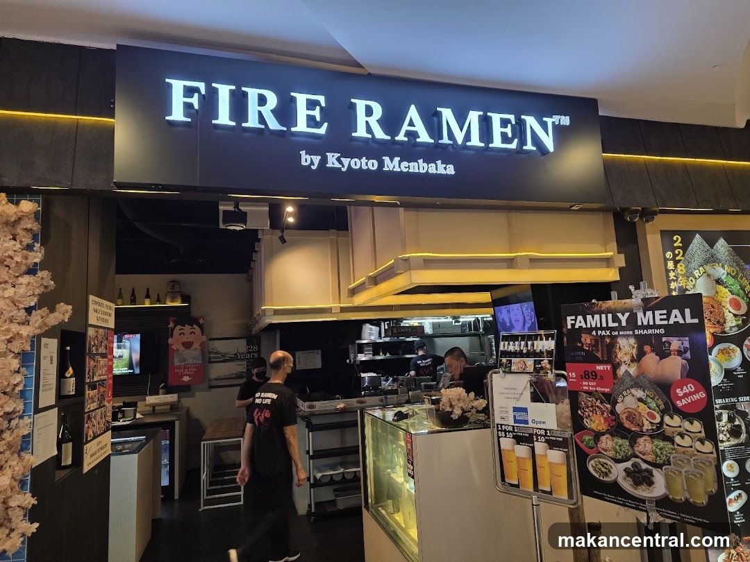 A brightly lit storefront with a large illuminated sign reading 'FIRE RAMEN™ by Kyoto Menbaka'. The entrance reveals the interior kitchen area where staff are working, and there is a display counter with a glass case. A decorative pink floral arrangement is on the left side of the frame.