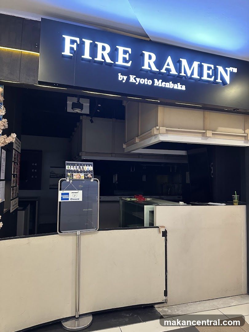 Exterior storefront with illuminated sign reading 'FIRE RAMEN™ by Kyoto Menbaka'. A closed sign is displayed on a stand in front of the counter. The entrance appears to be blocked or under maintenance, with no visible interior activity.