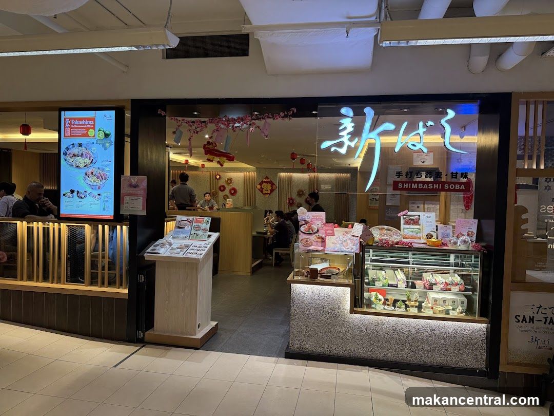 Storefront with illuminated Japanese characters '新ばし' and English text 'SHIMABASHI SOBA'. Glass window displays interior with patrons dining. Decor includes red lanterns and pink floral garlands. A display case shows food items. To the left, a digital menu board for Tokushima is mounted on a wooden railing.