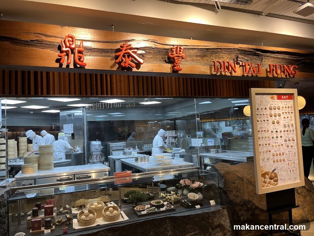 Storefront with wooden sign featuring red Chinese characters '鼎泰豐' and English name 'DIN TAI FUNG'. Glass window reveals kitchen area where staff in white uniforms prepare food. Display case below holds steamed dumplings, oysters, and other dishes. A stone-like base supports the display. To the right stands a menu board on an easel.