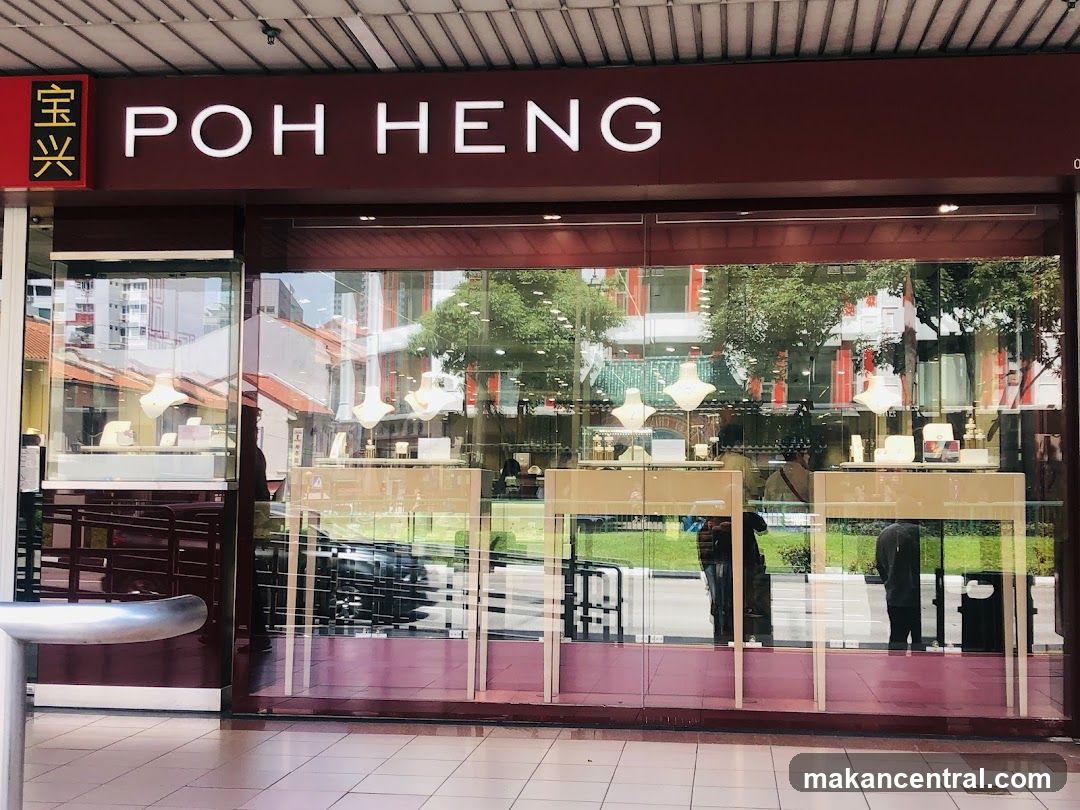 A storefront with a maroon sign displaying 'POH HENG' in white uppercase letters. To the left of the main sign is a vertical red box with Chinese characters '宝兴'. The shop has large glass windows reflecting the street, showing displays inside with jewelry on wooden tables under hanging lights. A metal bench is visible to the left outside the entrance.