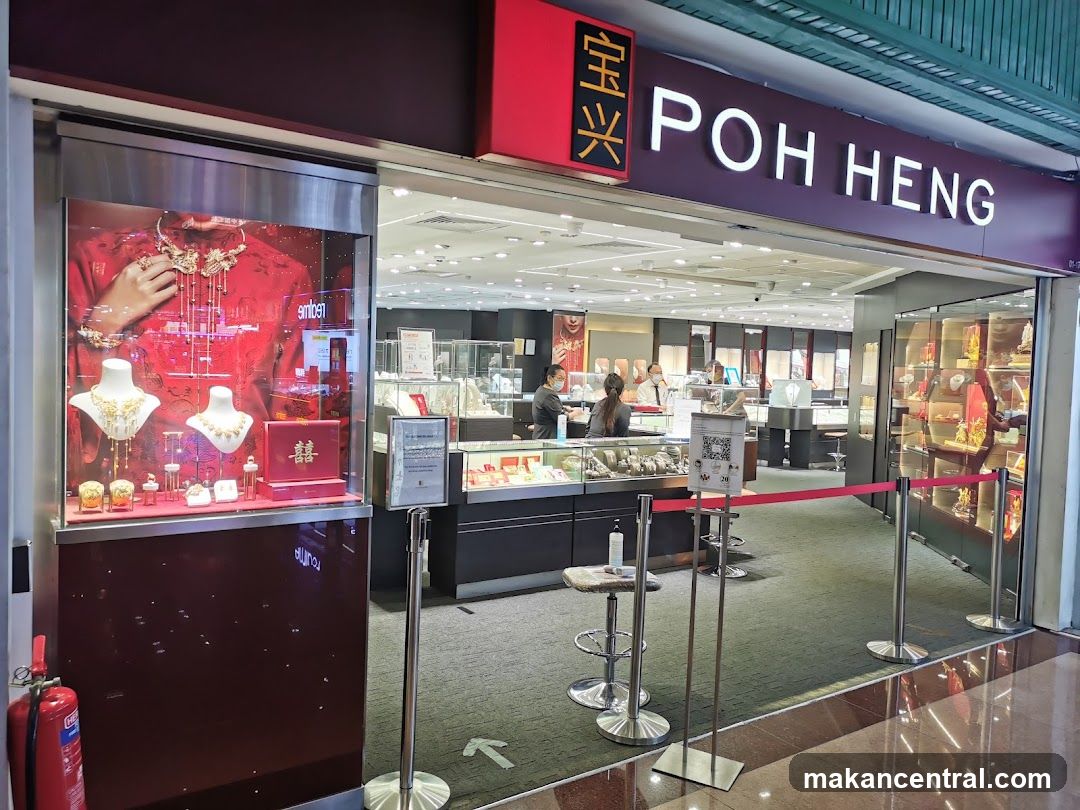 Storefront with prominent 'POH HENG' sign in white letters on a dark background, accompanied by Chinese characters '宝兴' on a red panel above the entrance. Glass display cases showcase jewelry, including necklaces and earrings on mannequins, with a red promotional backdrop featuring a bride's attire. Inside, customers are seen browsing products behind glass counters. Red velvet rope barriers guide foot traffic. A fire extinguisher is visible to the left.