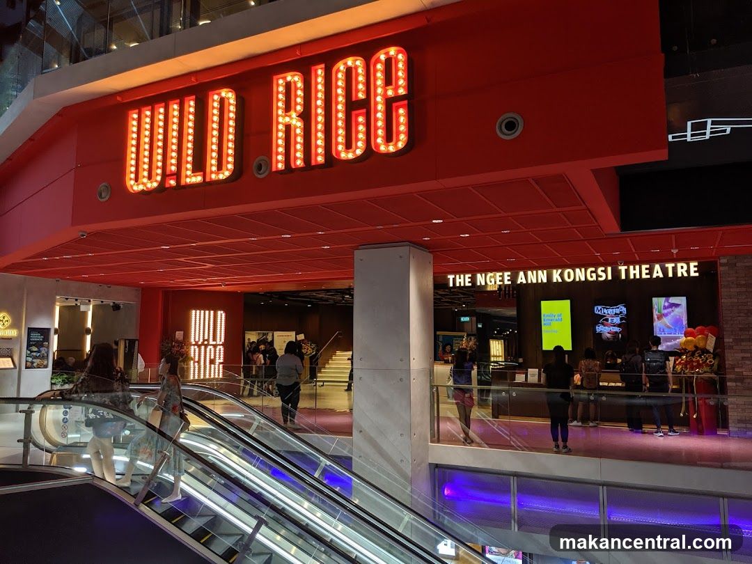Red illuminated sign with 'WILD RICE' in marquee letters above entrance. Below, 'THE NGEEN ANN KONGSI THEATRE' is displayed on a lit panel. Escalators lead to the level where the restaurant and theatre are located. People are seen walking or standing near the entrance.
