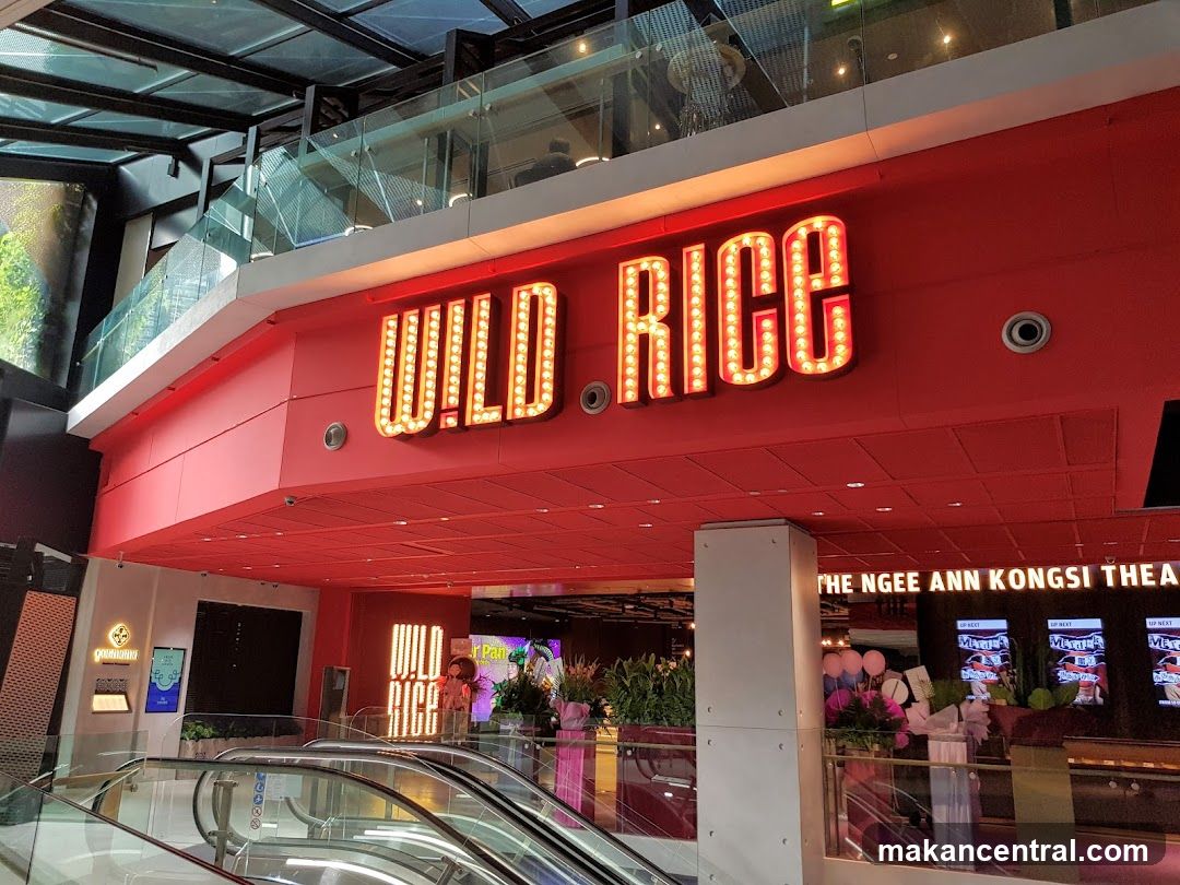 Interior view of a multi-level shopping mall with a prominent red neon sign reading 'WILD RICE' above the entrance. The area includes escalators leading to upper levels, glass railings, and potted plants near the entrance. To the right, signage indicates 'THE NGEES ANN KONGSI THEATRE'. No menu or printed materials are visible.