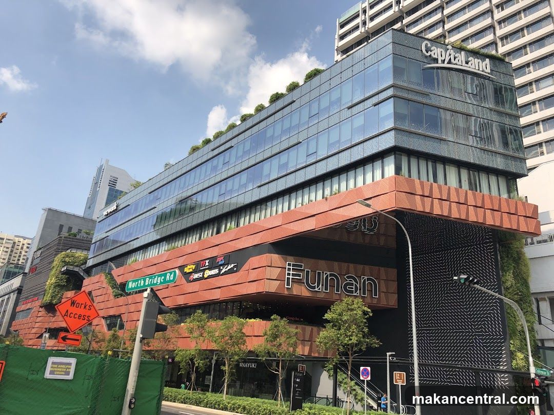 Modern multi-story building with glass facade and red brick lower section. 'Funan' sign prominently displayed on the front. Capitaland logo visible at top. Greenery integrated into design. Street signs indicate North Bridge Rd. Works Access sign and construction fencing present.