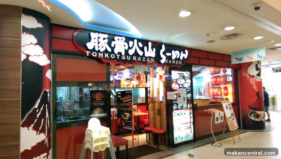 A brightly lit storefront with red and black signage featuring the name 'Tonkotsu Kazan Ramen' in both Japanese characters and English. The entrance has glass doors and windows showing interior seating, a white high chair outside, and promotional posters inside. A red pillar to the right depicts a ramen bowl. The ceiling above includes recessed lighting and a security camera.