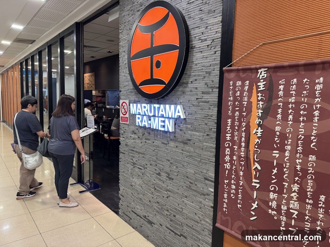 Storefront with gray brick wall featuring a circular orange logo above the illuminated "MARUTAMA RA-MEN" sign. Two customers stand at the entrance, facing inside. A red promotional banner with Japanese text hangs to the right.