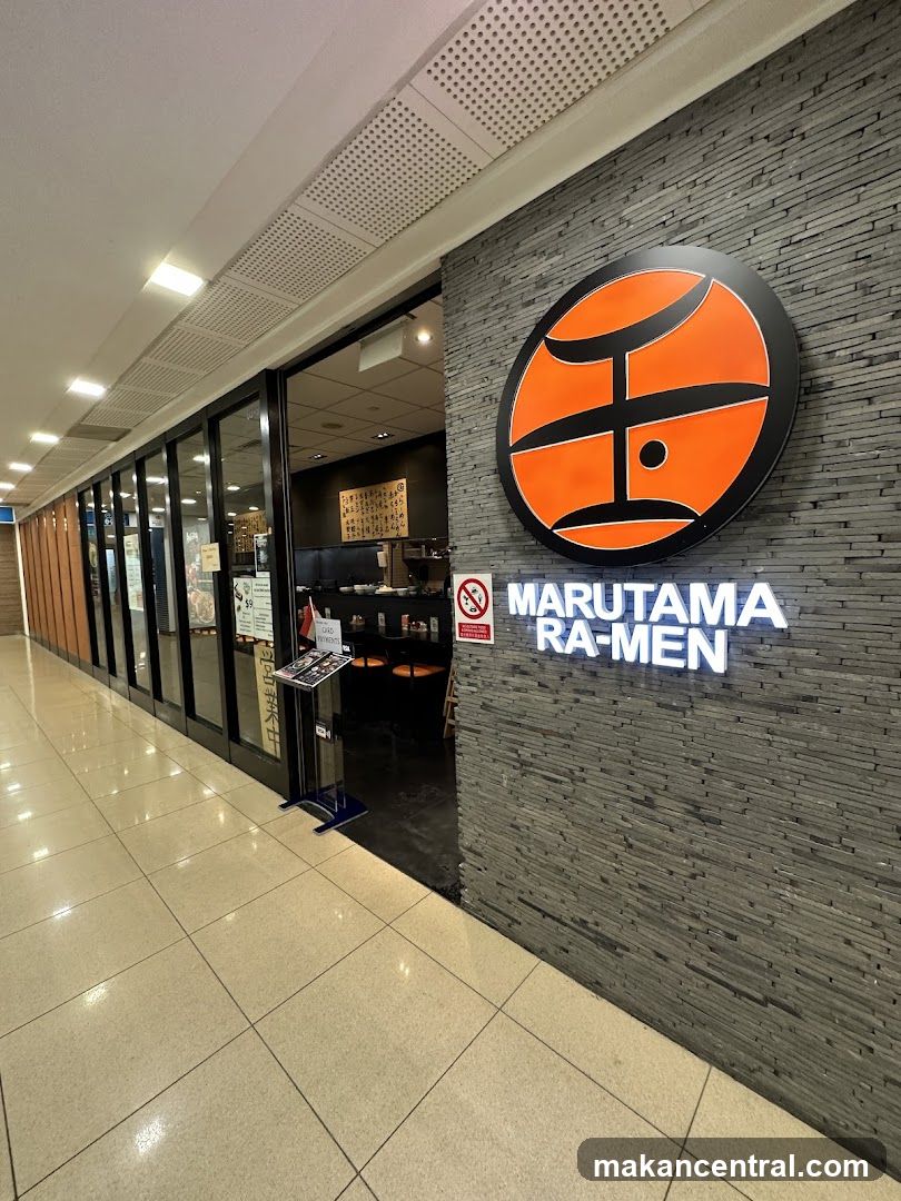 Storefront with a textured gray stone wall featuring an illuminated orange circular logo with black design elements above the glowing white "MARUTAMA RA-MEN" sign. Glass doors lead into the restaurant interior, which has dark ambient lighting and visible seating. A small stand holding printed materials is positioned near the entrance.