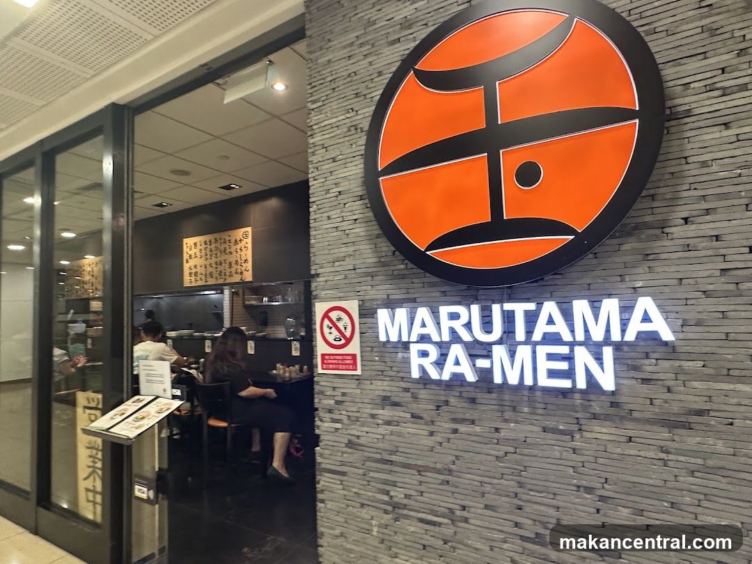 Exterior view of a ramen restaurant with a large orange circular logo on a gray brick wall. The name 'MARUTAMA RA-MEN' is illuminated in white letters below the logo. Glass doors lead into the dining area where patrons are seated. A no-smoking sign and menu stand are visible near the entrance.
