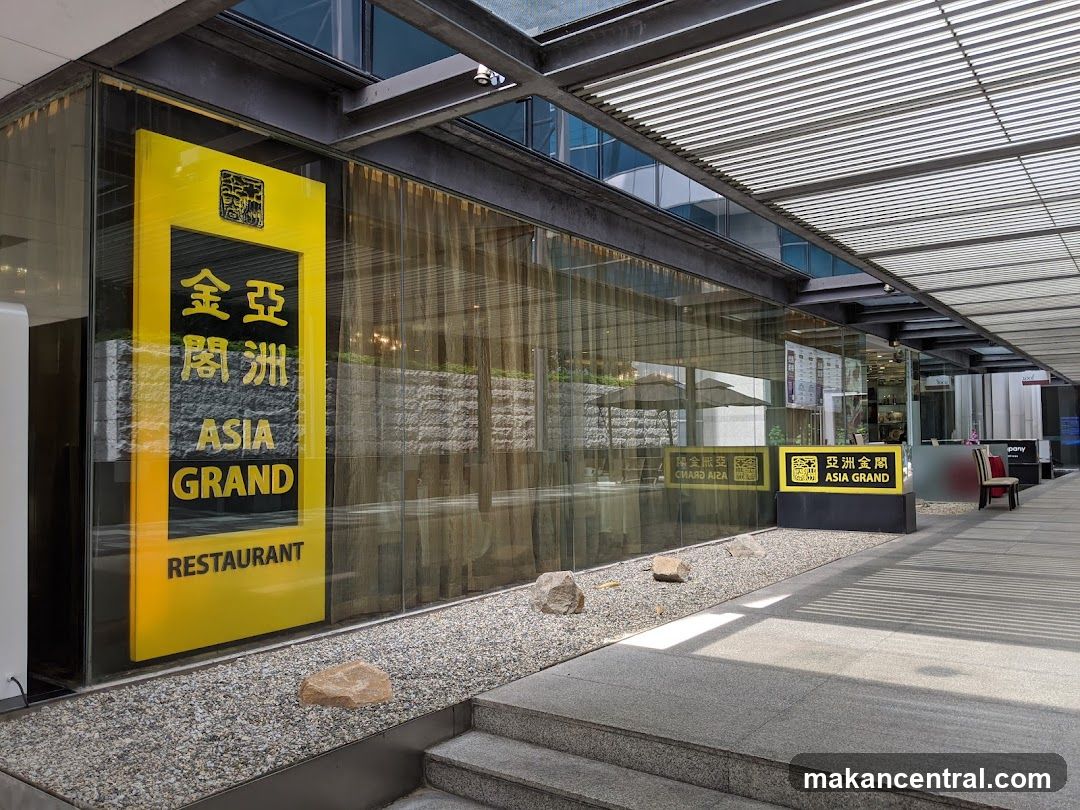Modern glass storefront with a bright yellow sign displaying "金亞洲閣" vertically above "ASIA GRAND RESTAURANT" in English. The entrance features a gravel garden bed with rocks, leading to a covered walkway with slatted roof. Reflections of interior lighting and umbrellas are visible through the glass. A smaller version of the logo appears on a black base near the sidewalk.