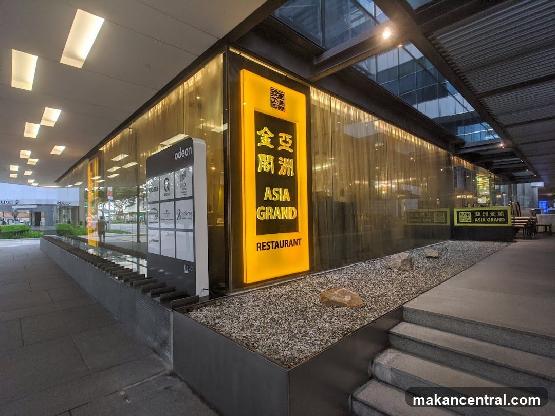 Modern storefront with illuminated yellow sign displaying "亞洲金閣" vertically and "ASIA GRAND RESTAURANT" below it. The facade features glass panels reflecting interior lights. A low gravel garden bed with stones borders the entrance, leading to stone steps. Adjacent signage includes a white panel with logos of Odeon and other brands. Background shows urban architecture and a pedestrian pathway.