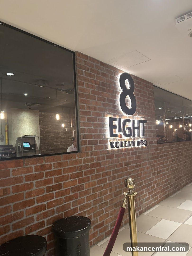 Brick wall facade with illuminated sign displaying '8 EIGHT KOREAN BBQ'. Glass windows reflect interior lighting and a staff member. A gold stanchion with red velvet rope stands in front of the entrance.