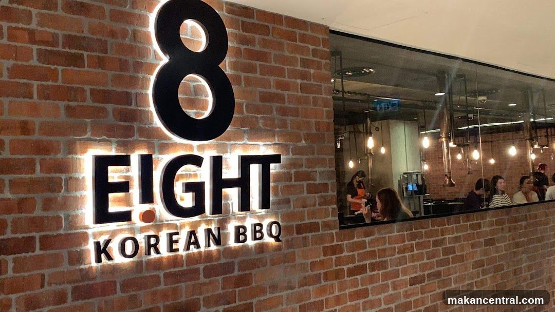 Exterior storefront with exposed brick wall featuring illuminated black signage reading '8 EIGHT KOREAN BBQ'. A large glass window reveals the interior dining area where patrons are seated and staff are working. The lighting inside includes hanging bulbs, and the ambiance appears modern and casual.