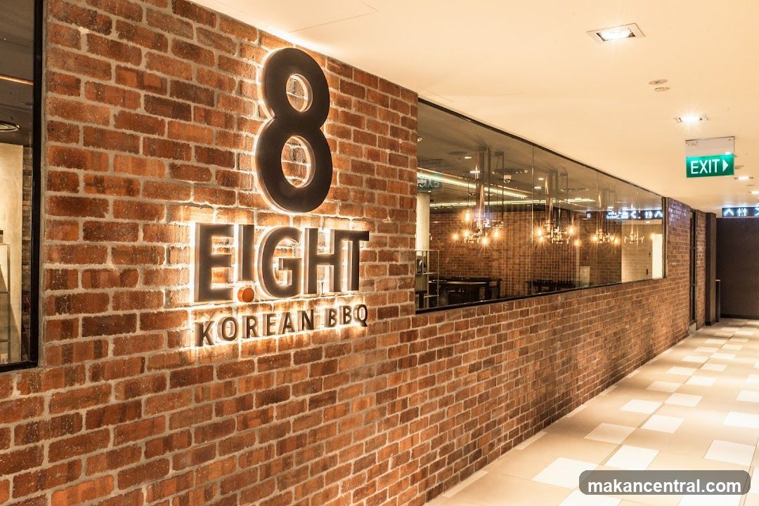 Exterior storefront with exposed brick wall featuring illuminated signage reading '8 EIGHT KOREAN BBQ'. Large glass window reveals interior dining area with chandeliers and tables. Floor is tiled in a checkered pattern. Green 'EXIT' sign visible overhead.