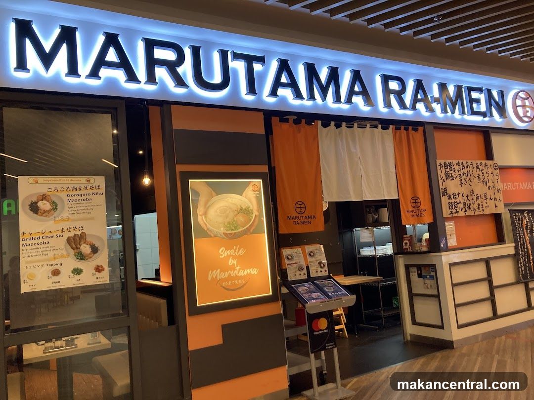 Brightly lit sign with 'MARUTAMA RAMEN' in large illuminated letters above the entrance. Orange and black facade with hanging fabric curtains. Visible interior includes counter, seating area, and a standing payment kiosk. Glass door displays printed menu boards on left side.
