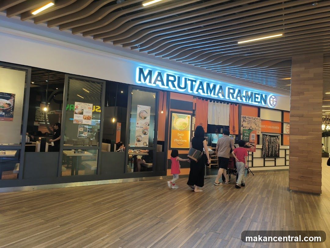 Brightly lit neon sign reading 'MARUTAMA RA-MEN' above the entrance. Large glass windows display interior seating and staff. A family of three walks toward the entrance. Modern wooden flooring and ceiling with wavy slats. Orange and white decor inside.