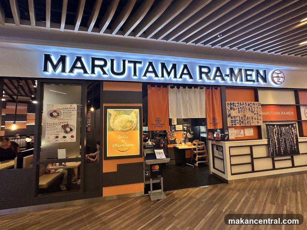 Brightly lit sign with 'MARUTAMA RA-MEN' in large letters above the entrance. The facade features orange and black accents, a wooden floor, and a ceiling with slatted panels. Visible inside are tables, chairs, and a counter area. A promotional poster is mounted on the wall to the left of the entrance.
