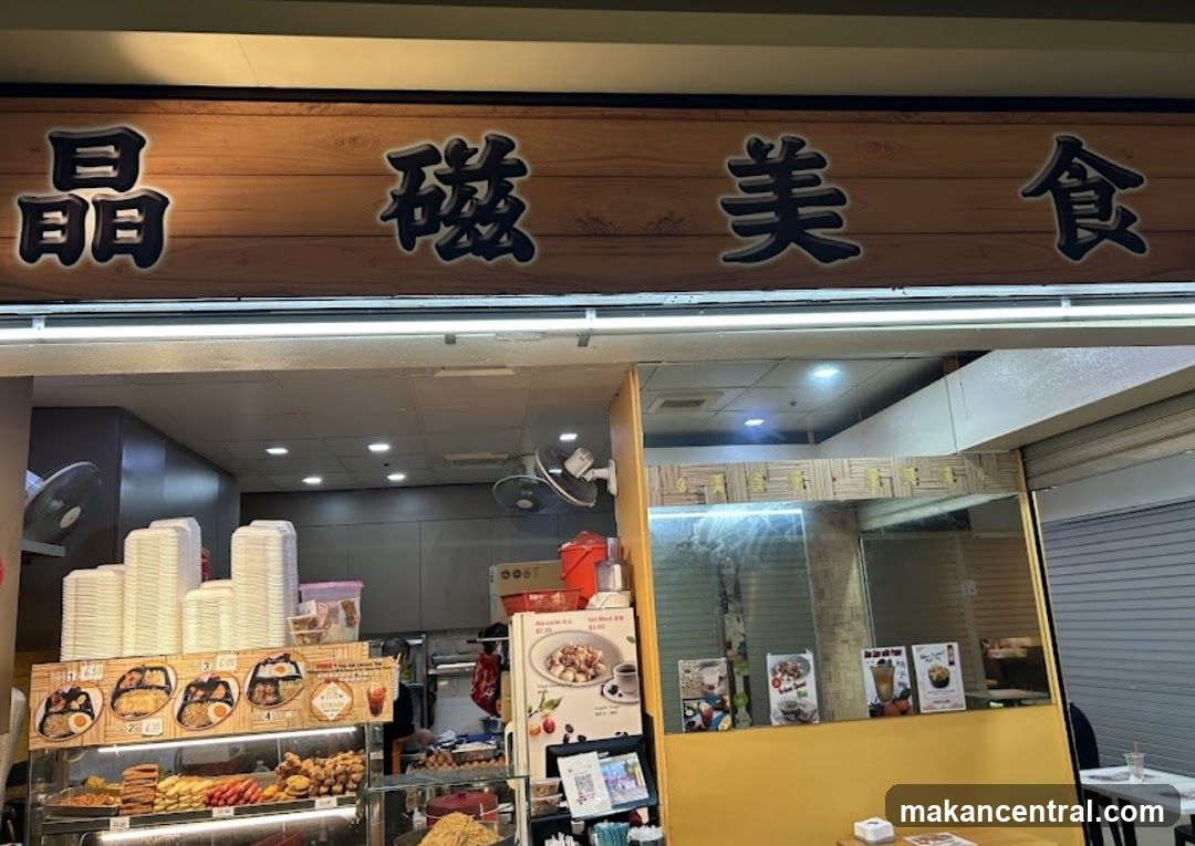 A storefront with a wooden signboard displaying the characters '晶磁美食' in black outline against a light wood background. Below the sign, the interior of the shop is partially visible, showing stacked disposable plates, a display case with food items, and a counter area with equipment like fans and refrigeration units. The entrance appears open to the public, revealing part of the kitchen and seating area.