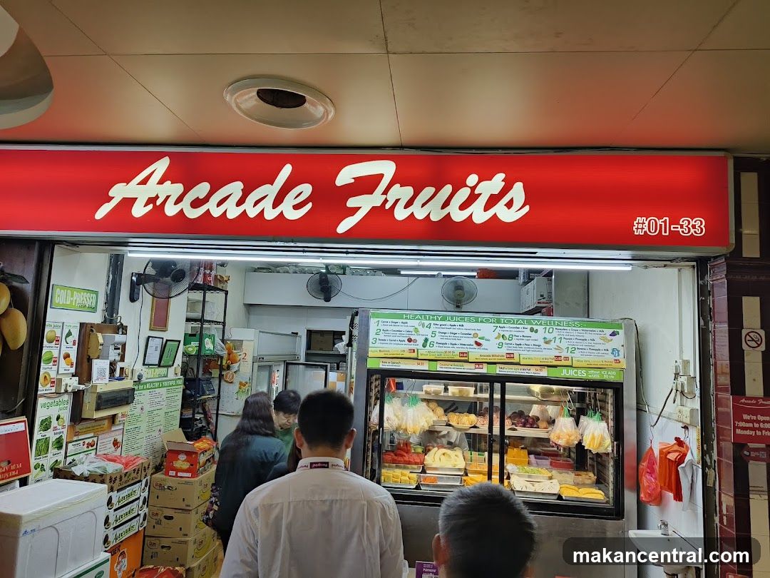 Storefront with red sign displaying 'Arcade Fruits #01-33'. Interior shows refrigerated display of fruits and juices, customers inside, shelves with boxes, fans on walls, and a sink to the right.