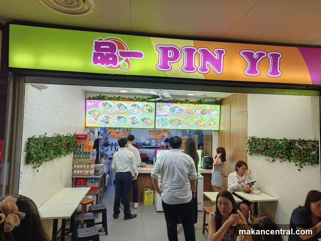 A brightly lit storefront with a green and orange sign displaying '品一 PIN YI' in stylized lettering. The entrance reveals the interior where customers are ordering food. Above the counter, there is a digital menu board showing images of dishes. Greenery decorates the walls on either side.