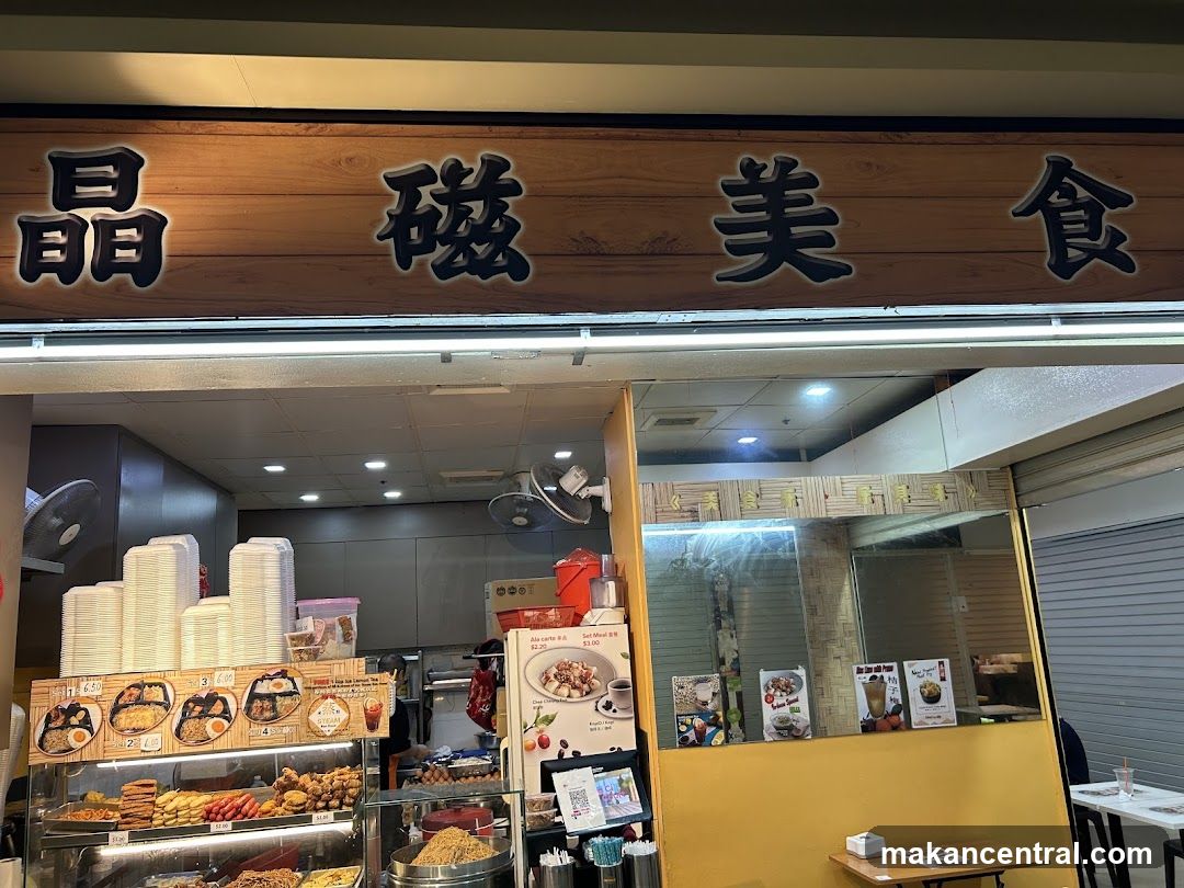 A storefront with a wooden sign bearing the characters '晶磁美食' in black calligraphy. The shop appears to be an eatery with a display case filled with food items like pastries and fried snacks. Stacks of white disposable containers are visible on the left, and a fan is mounted above the counter area. A yellow wall and glass partition separate the interior from the entrance. No menu board or printed material is clearly visible on the front facade.