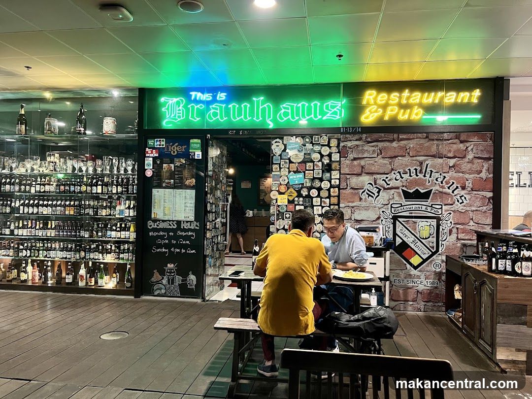 Neon sign reads 'This is Brauhaus Restaurant & Pub' above the entrance. Brick wall with logo and emblem featuring a crown and beer mug. Glass display shelves filled with bottles on left side. Two patrons seated at wooden table in foreground, facing away from camera. Business hours posted on blackboard door.