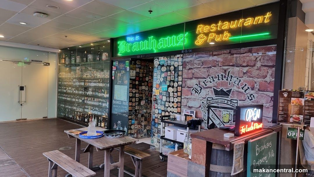 Exterior storefront with neon green and yellow signage reading 'This is Brauhaus Restaurant & Pub'. The facade features a brick wall design with the logo and name painted on it, along with an illuminated 'OPEN' sign. A glass display case holds numerous beer bottles behind the counter area. Wooden picnic-style tables and benches are positioned outside the entrance.
