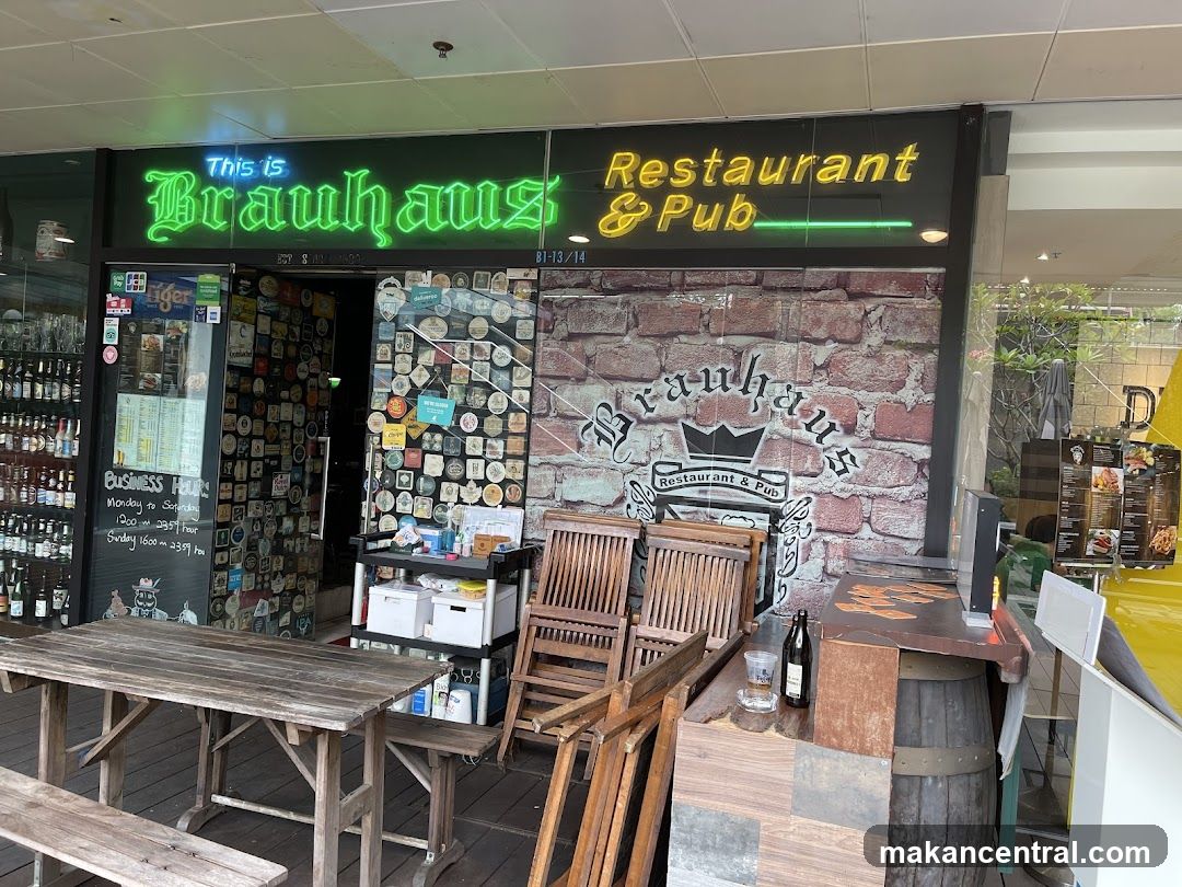 Exterior storefront with neon sign reading 'This is Brauhaus Restaurant & Pub'. The facade features a brick wall design with the logo and name painted on it. Outside seating includes wooden tables and chairs. A glass door displays business hours and beer logos, while shelves inside show bottles. A wooden barrel sits near the counter.