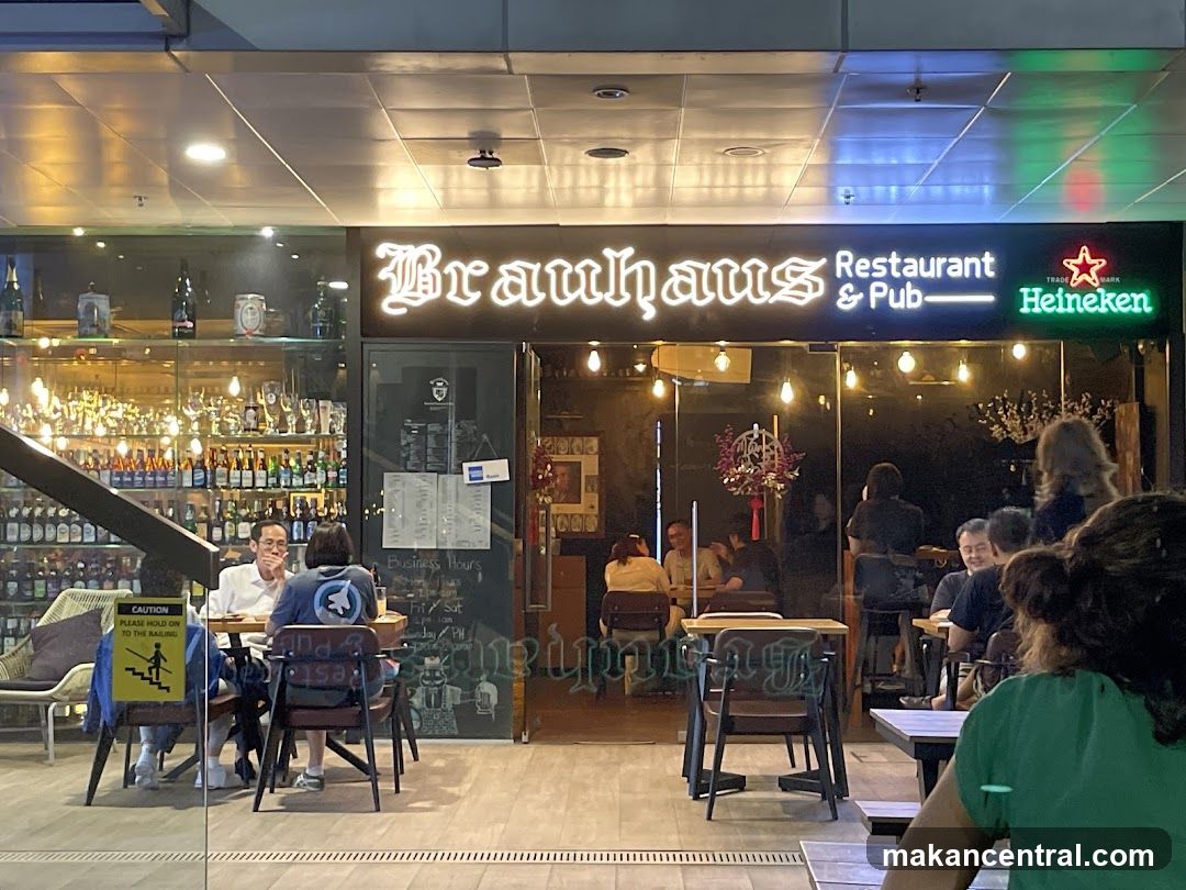 Exterior storefront with illuminated sign reading 'Brauhaus Restaurant & Pub' and a Heineken logo to the right. Glass windows display shelves of bottles inside; patrons are seated at tables outside and inside. A yellow caution sign is on the left. The entrance has a dark interior with warm lighting, and a menu board is visible on the glass door.