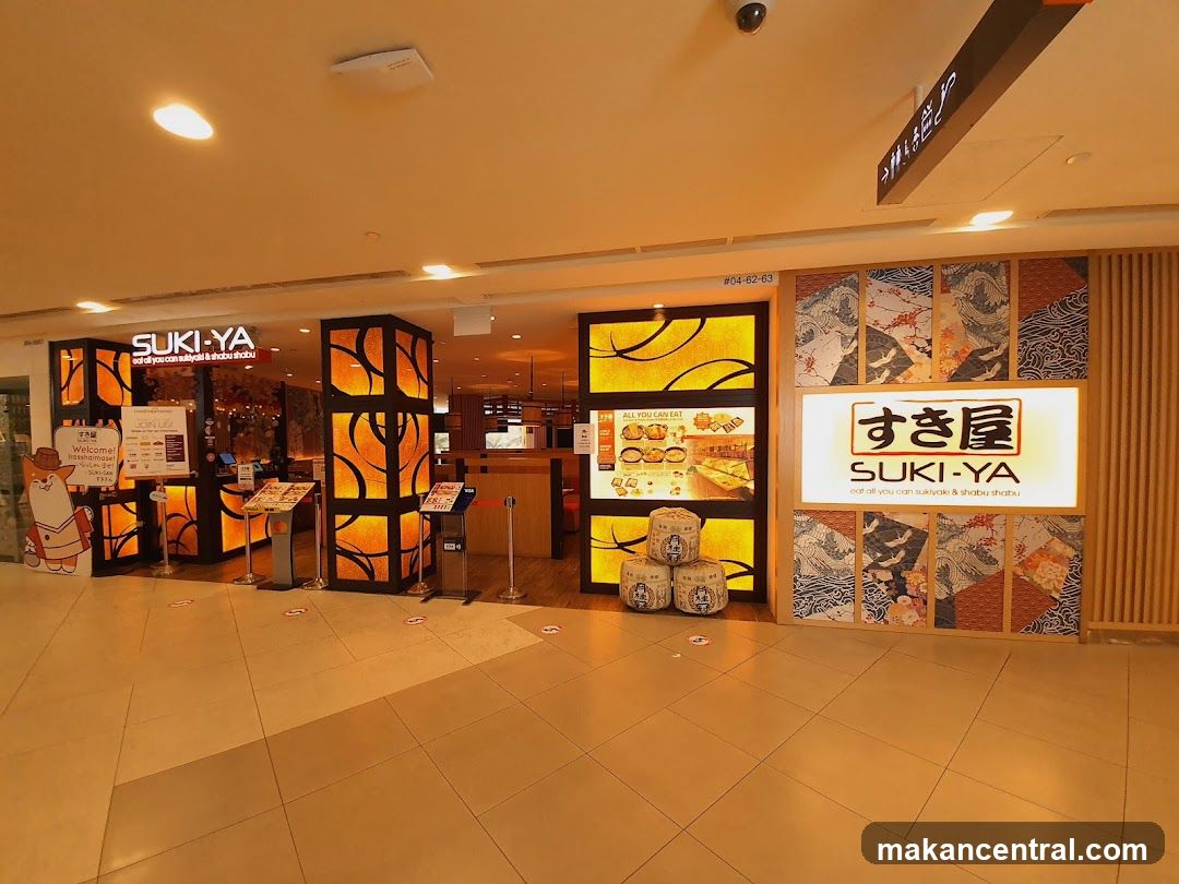 The storefront features illuminated signage with the name 'SUKI-YA' in English and Japanese characters. The entrance is framed by decorative orange-lit panels with circular patterns. A large digital menu board displays 'ALL YOU CAN EAT' options, including images of food items. Three stacked barrels sit near the entrance. The interior is warmly lit, showing a counter area and seating. A cartoon mascot figure stands to the left. Above the entrance, a sign reads '#04-82-63'.