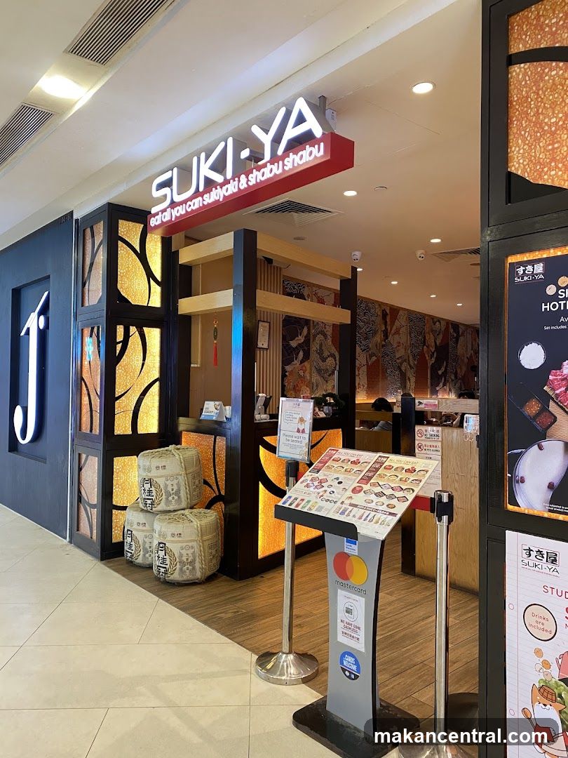 Storefront with illuminated sign reading 'SUKI-YA' and subtext 'eat all you can sukiyaki & shabu shabu'. Entrance features decorative glass panels with golden patterns, wooden accents, and stacked rice barrels outside the door. A menu stand with Mastercard logo is positioned at the entrance.