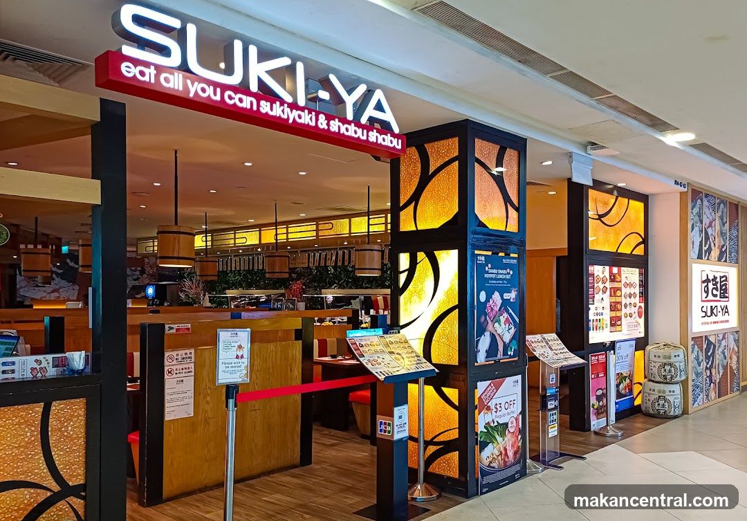 The storefront features a bright red LED sign with 'SUKI-YA' in large white letters above the entrance. Below it reads 'eat all you can sukiyaki & shabu shabu'. The entrance is framed by black pillars with illuminated orange and yellow patterned panels. Inside, wooden tables and chairs are visible under warm lighting. A red velvet rope barrier stands at the entrance. To the right, promotional posters and advertisements are displayed on easels.