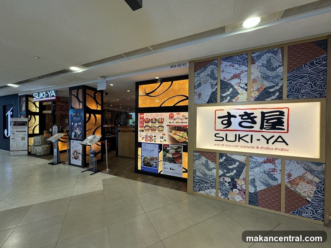 Storefront facade with illuminated sign displaying 'すき屋 SUKI·YA' and tagline 'eat all you can sukiyaki & shabu shabu'. Background wall features traditional Japanese wave and floral patterned panels. Above entrance, unit number #04-62-63 is visible. Interior lighting and promotional posters are partially visible inside the shop.