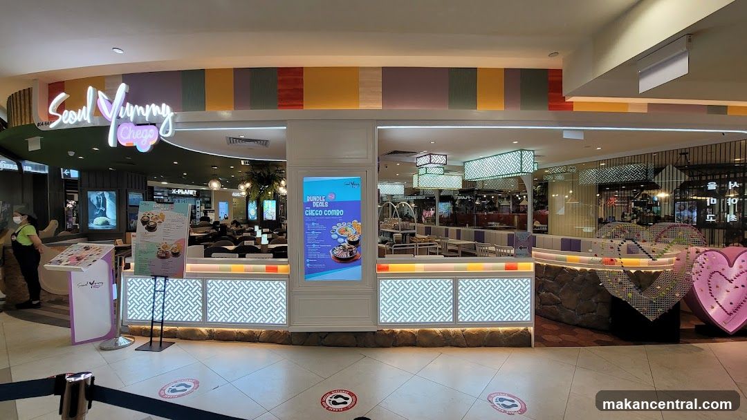 Storefront with illuminated sign 'Seoul Yummy Cheeze' above entrance; colorful striped ceiling paneling; glass windows showing interior seating area; counter with patterned lighting panels; staff in green shirt near entrance; floor has social distancing markers.