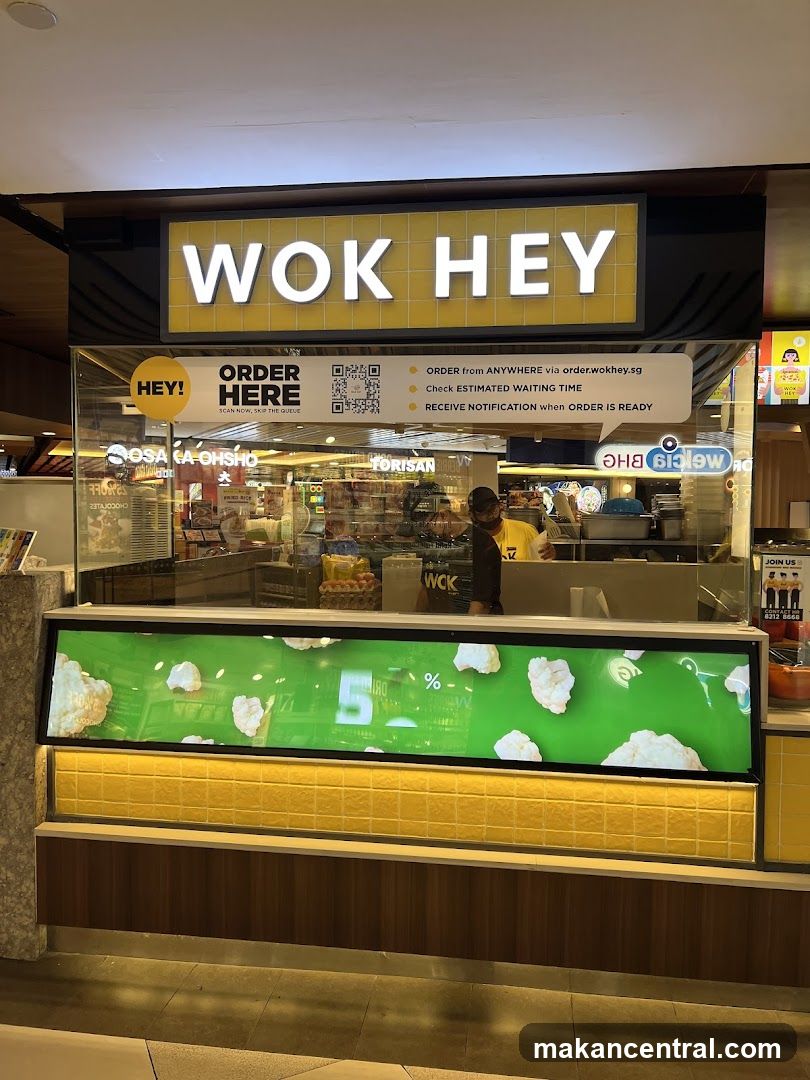 A brightly lit food stall with a prominent yellow sign displaying 'WOK HEY' in white letters. The counter is enclosed by glass, showing staff inside the kitchen area. A digital display below shows promotional graphics with popcorn imagery and a '5%' discount indicator. Above the counter, there are instructions for ordering via QR code and website, along with features like estimated waiting time and order readiness notifications.