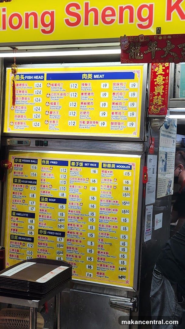 Yellow menu board with Chinese and English text listing food categories: Fish Head, Meat, Prawn Ball, Fried, Set Rice, Noodles, Vegetables, Soup, Omelette, Beancurd, Fried Rice. Prices in dollars. Menu displayed on a stall counter.