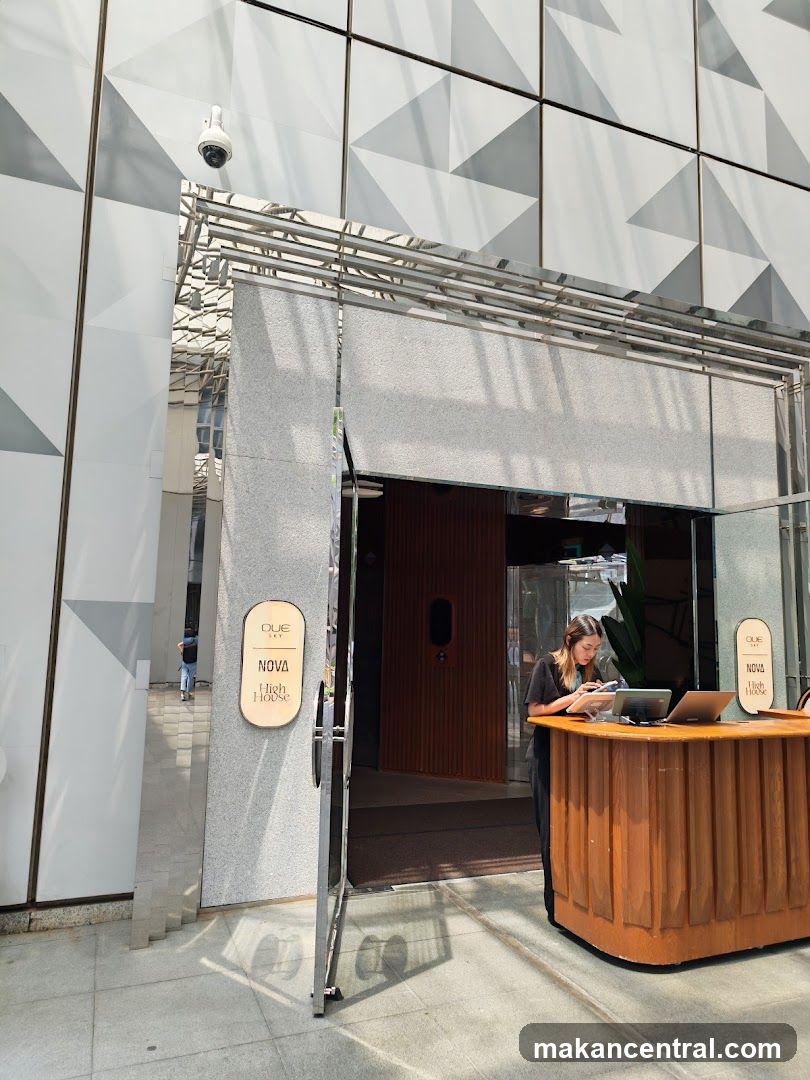 Modern building facade with geometric white and gray panels. A glass door opens into a lobby with a wooden reception counter where an employee is seated. Two oval signs on the pillars read 'NOVA High House'. A security camera is mounted above the entrance.