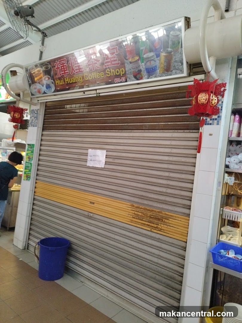 Closed storefront with metal roller shutter door, yellow stripe, red lantern hanging on right side, blue bucket beside entrance, signage above displaying shop name in Chinese and English with product imagery, person working at adjacent stall to left.