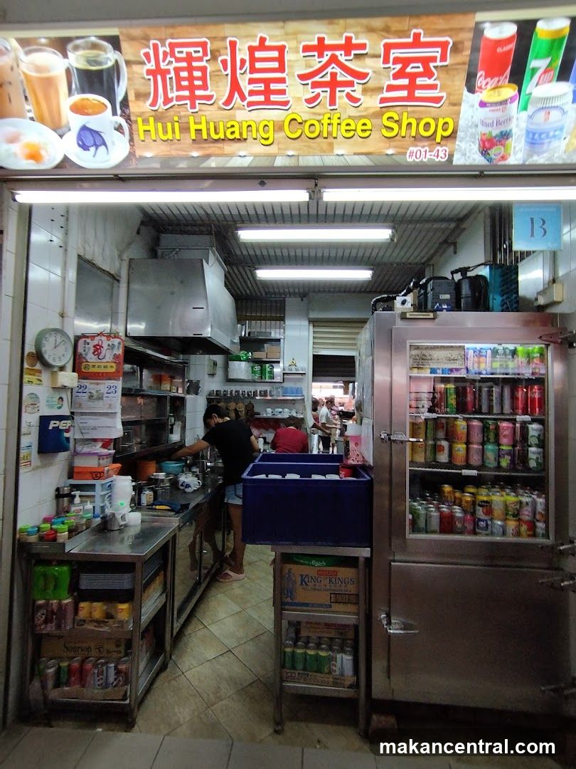 Storefront with signboard displaying "輝煌茶室" and "Hui Huang Coffee Shop #01-43" above entrance. Interior view shows kitchen area, staff working, refrigerated beverage display, and tiled floor. Signage includes product images (coffee cups), soda cans (Coca-Cola, 7UP) on right side of sign. No menu board or printed material visible.