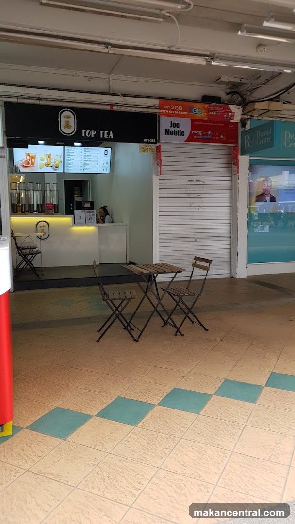 Exterior view of a tea shop with 'TOP TEA' sign above counter. A staff member is visible behind the counter. Adjacent to it, there's a shuttered 'Joe Mobile' store and a dental center sign. Two foldable tables and chairs are placed outside on tiled floor.