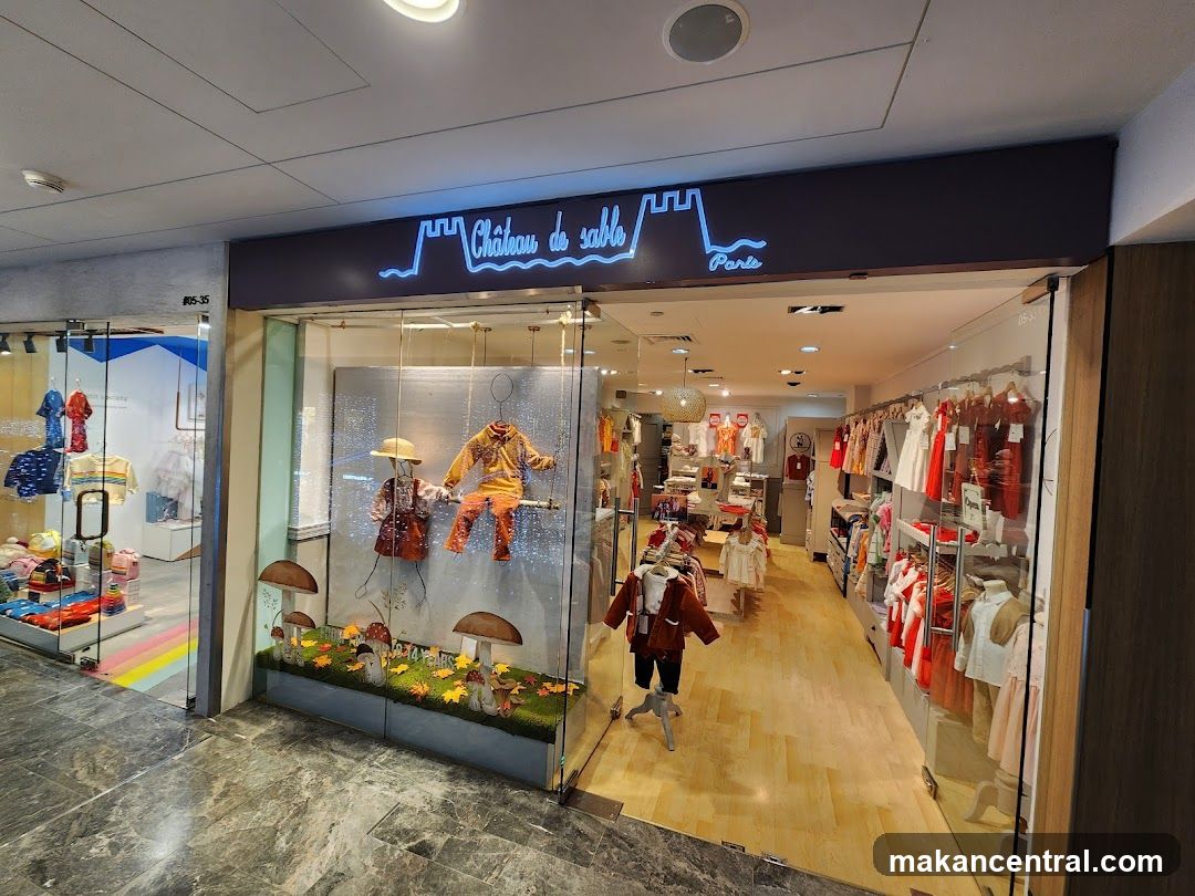 Storefront with neon sign displaying 'Château de sable Paris' above glass entrance. Display window features a fantasy-themed setup with mannequins in costumes, mushrooms, and autumn leaves. Interior shows wooden floors, racks of children's clothing, and bright lighting. Shop is located within a mall environment.