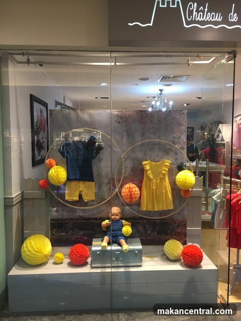 Glass display window with neon sign 'Château de' above entrance. Interior features a baby doll seated on a silver suitcase surrounded by yellow and red paper lanterns. Behind the doll, two circular hoops hold children's clothing: a blue shirt and shorts on one side, a yellow dress on the other. A textured gray backdrop is visible behind the displays. Reflections of store interior and shoppers are seen in glass. The shop appears to be a children’s clothing store.