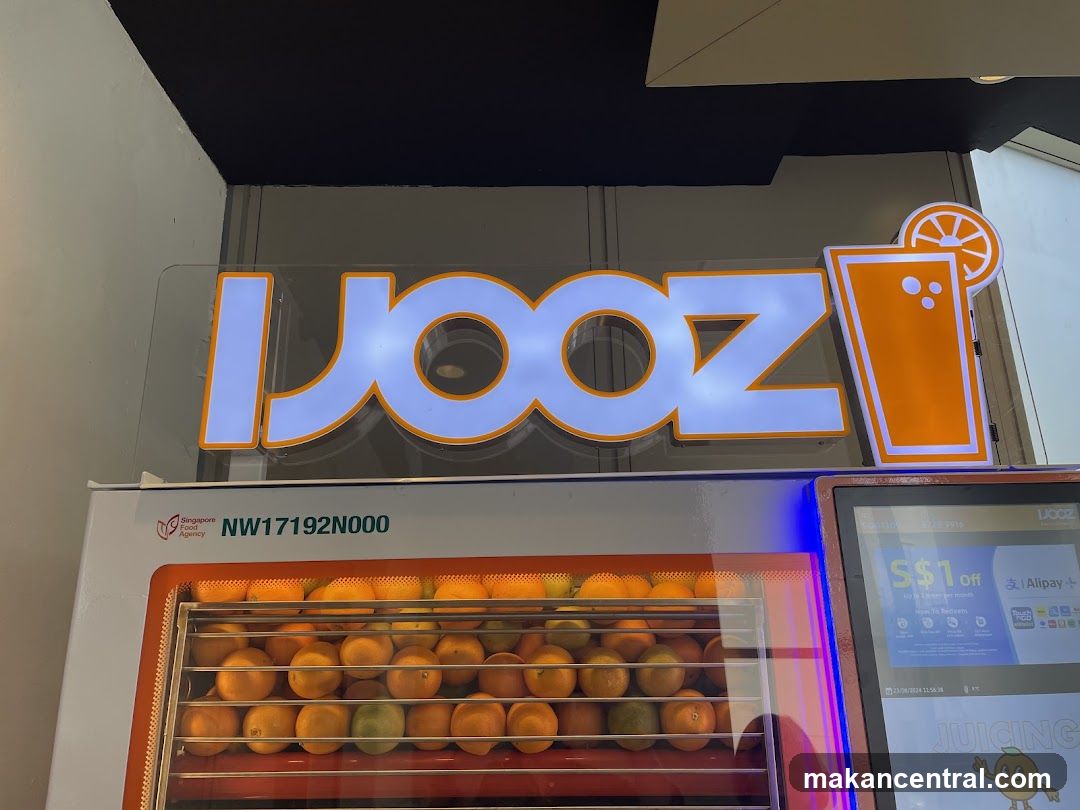 Brightly lit neon sign with the word 'JOOZ' in white letters outlined in orange, accompanied by a stylized orange juice glass icon. Below is a refrigerated display unit filled with oranges and limes, bearing the Singapore Food Agency registration number NW17192N000. To the right, an illuminated touchscreen kiosk displays promotional offers including '$1 Off' via Alipay+ and mentions 'JUICING'. The setup suggests a modern, automated juice vending machine located indoors or within a commercial space.