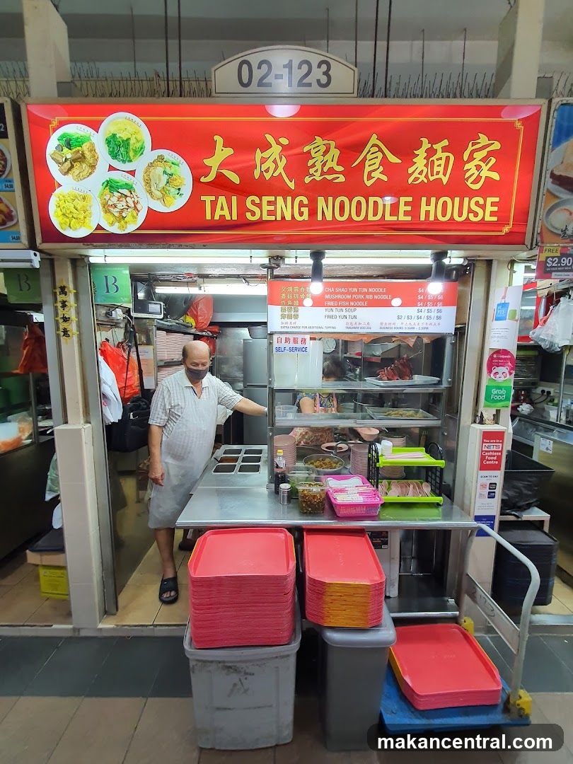 Red signboard with yellow Chinese characters '大成熟食麵家' and English name 'TAI SENG NOODLE HOUSE'. Shop number 02-123 above entrance. Interior shows a chef in mask, stainless steel counter, stacked red trays, and glass display cases with food. Self-service label visible.